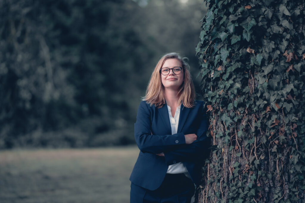 Woman in navy blazer and white shirt stands beside a ivy-covered wall outdoors, arms crossed, smiling softly.
